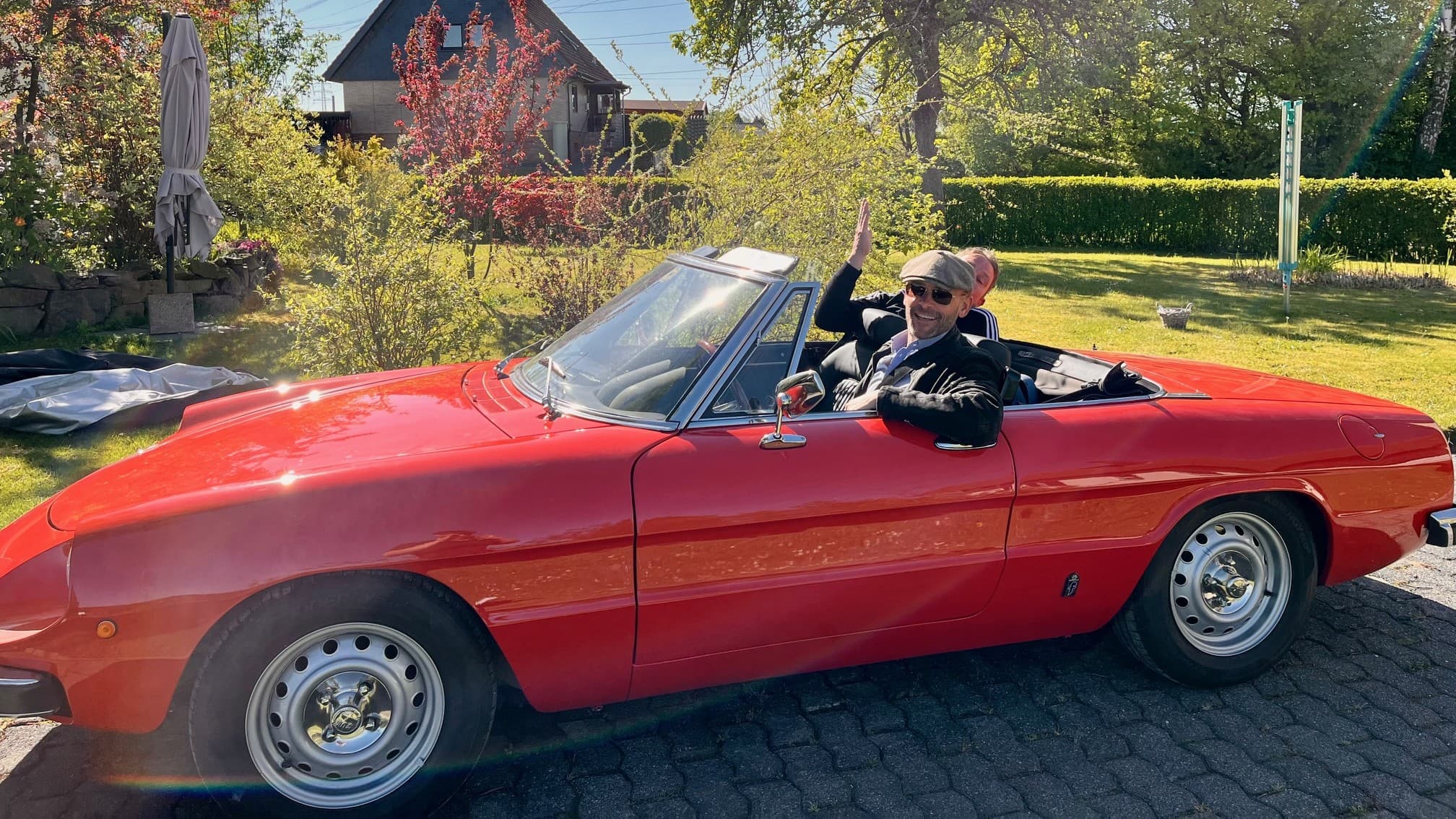 Sean Kollak im roten Alfa Romeo Spider Baujahr 1977 auf Kopfsteinpflaster – winkend, Sonnenbrille, Schiebermütze – Metapher für das Innehalten, bevor man wegdämmert