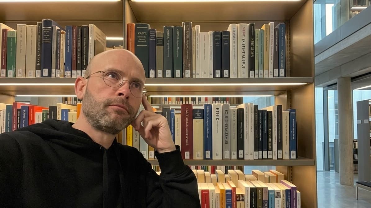 Sean Kollak nachdenklich vor Philosophie-Regal – implizites Wissen lässt sich nicht aufschreiben
