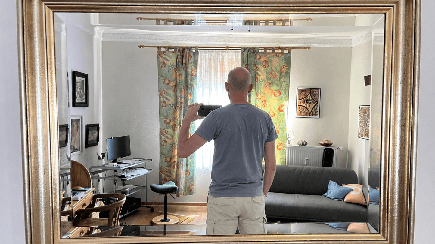 Sean Kollak fotografiert sich in einem goldgerahmten Spiegel – Rücken zur Kamera, Smartphone erhoben, Schreibtisch und Sofa im Hintergrund