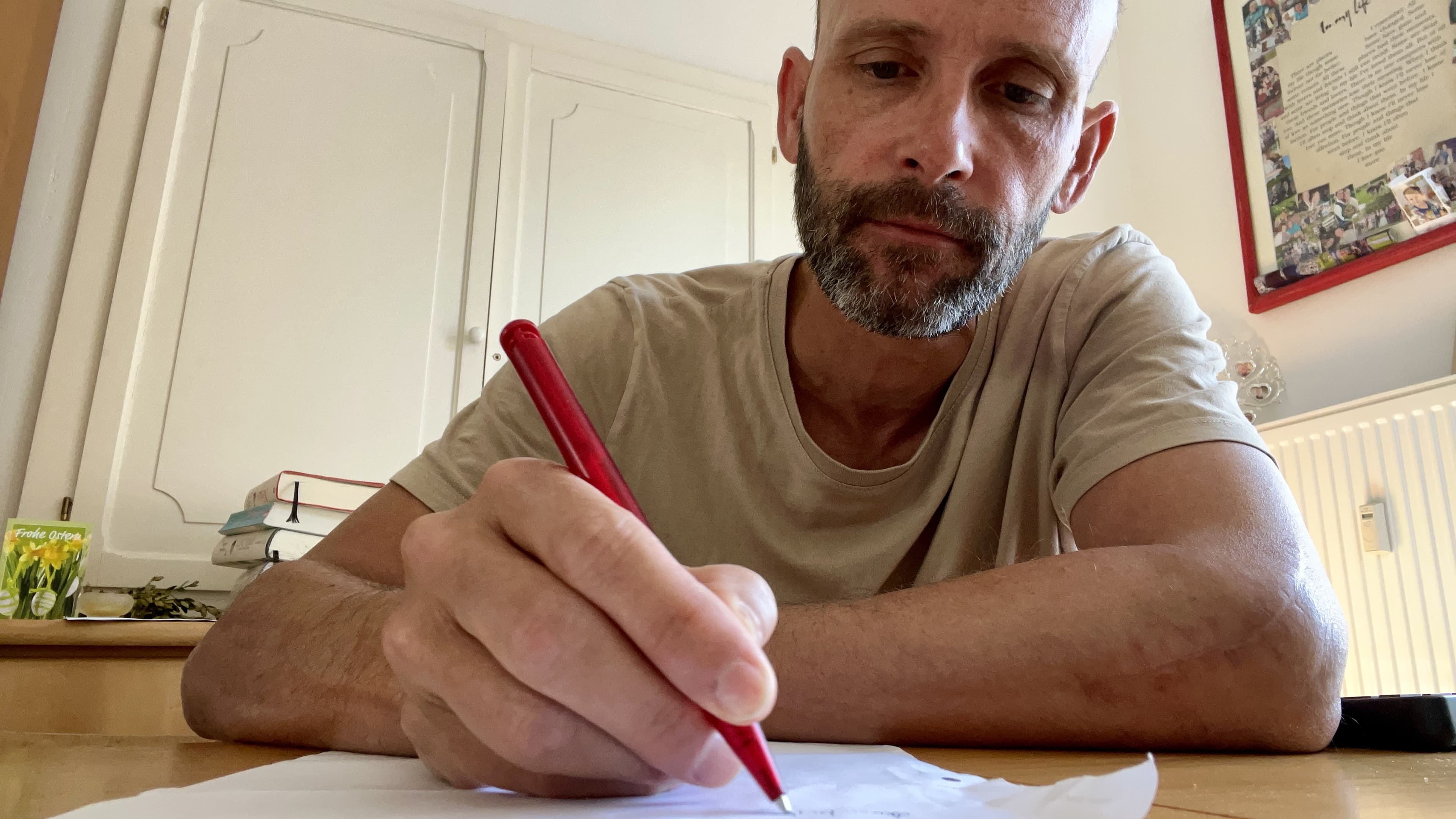 Sean Kollak schreibt mit rotem Stift einen Brief – konzentrierter Blick, Holztisch, Bücher im Hintergrund. Ein Foto als Beweis: Jemand war da.