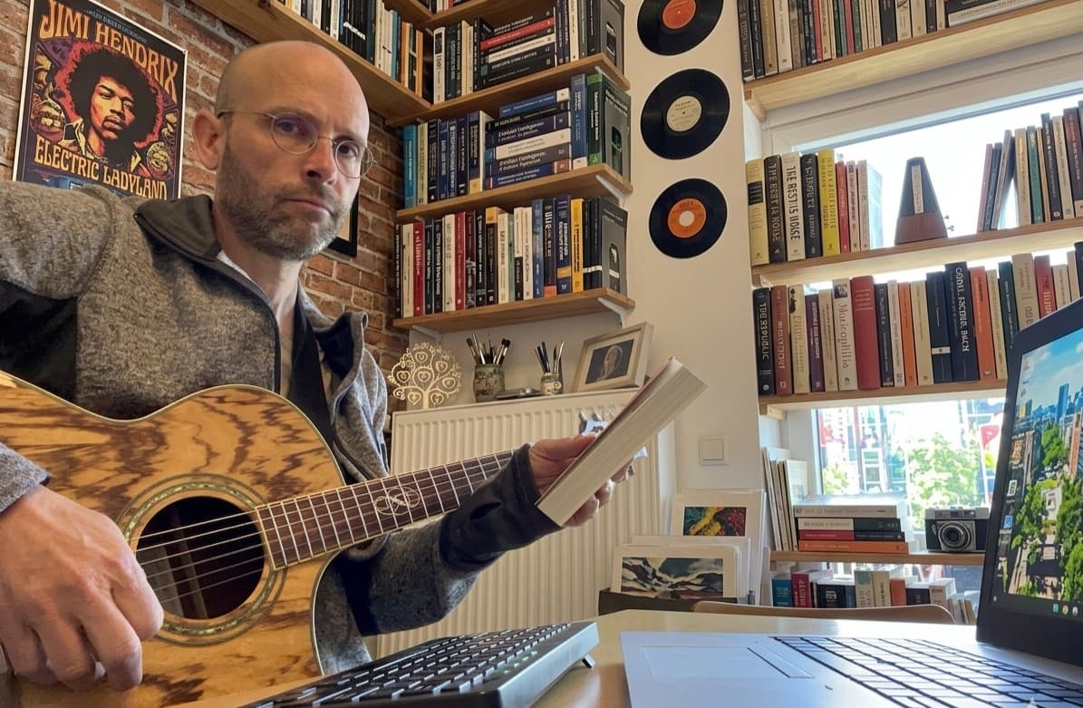 Sean Kollak sitzt mit Akustikgitarre und aufgeschlagenem Buch in seinem Arbeitszimmer – Bücherregale mit Schallplatten und Jimi-Hendrix-Poster im Hintergrund