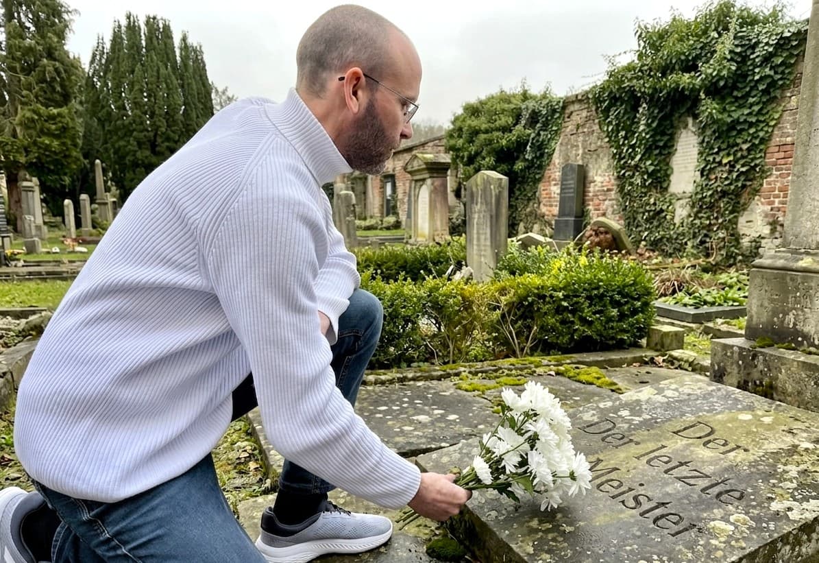 Sean Kollak legt weiße Blumen auf einen verwitterten Grabstein mit der Inschrift Der letzte Meister – Wissens-Entropie im deutschen Mittelstand