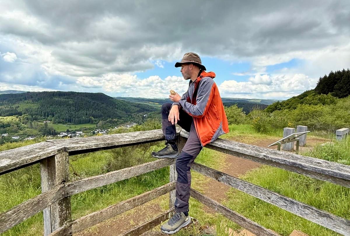 Sean Kollak sitzt auf einem Holzzaun am Eifelsteig und schaut in die hügelige Waldlandschaft der Eifel – auf dem Weg zwischen Denken und Spüren