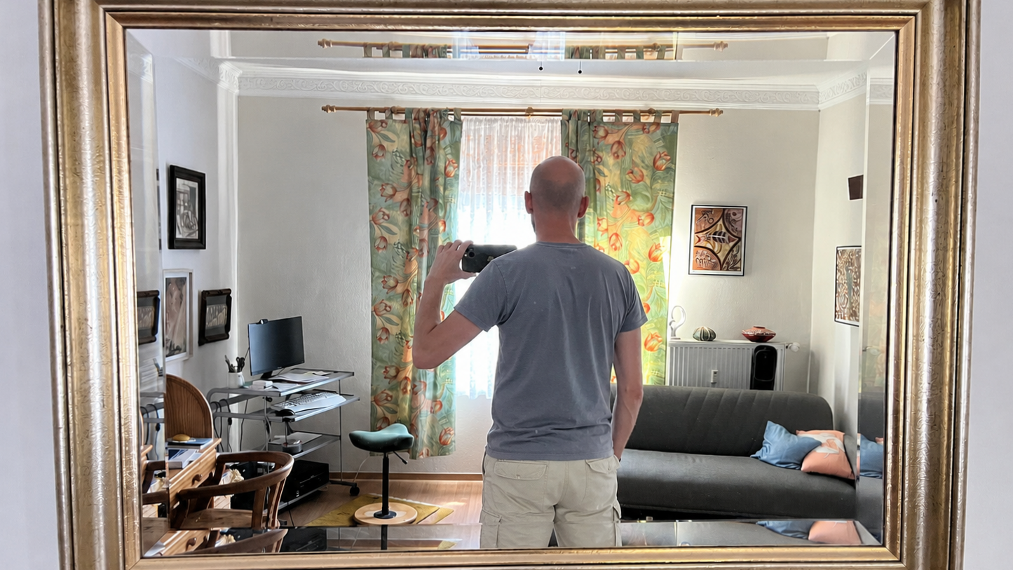 Sean Kollak fotografiert sich in einem goldgerahmten Spiegel – Rücken zur Kamera, Smartphone erhoben, Schreibtisch und Sofa im Hintergrund